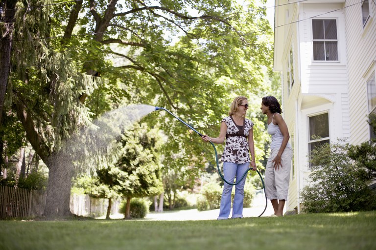 Two women chatting in house backyard, one watering lawn with hose