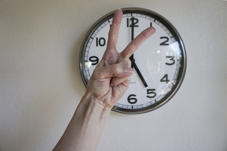 Hand showing victory sign in front of a clock, at five p.m.